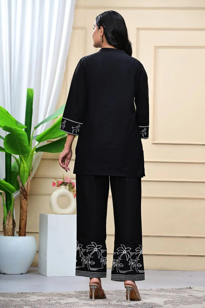 Person wearing a black outfit with white embroidery in a minimal indoor setting.